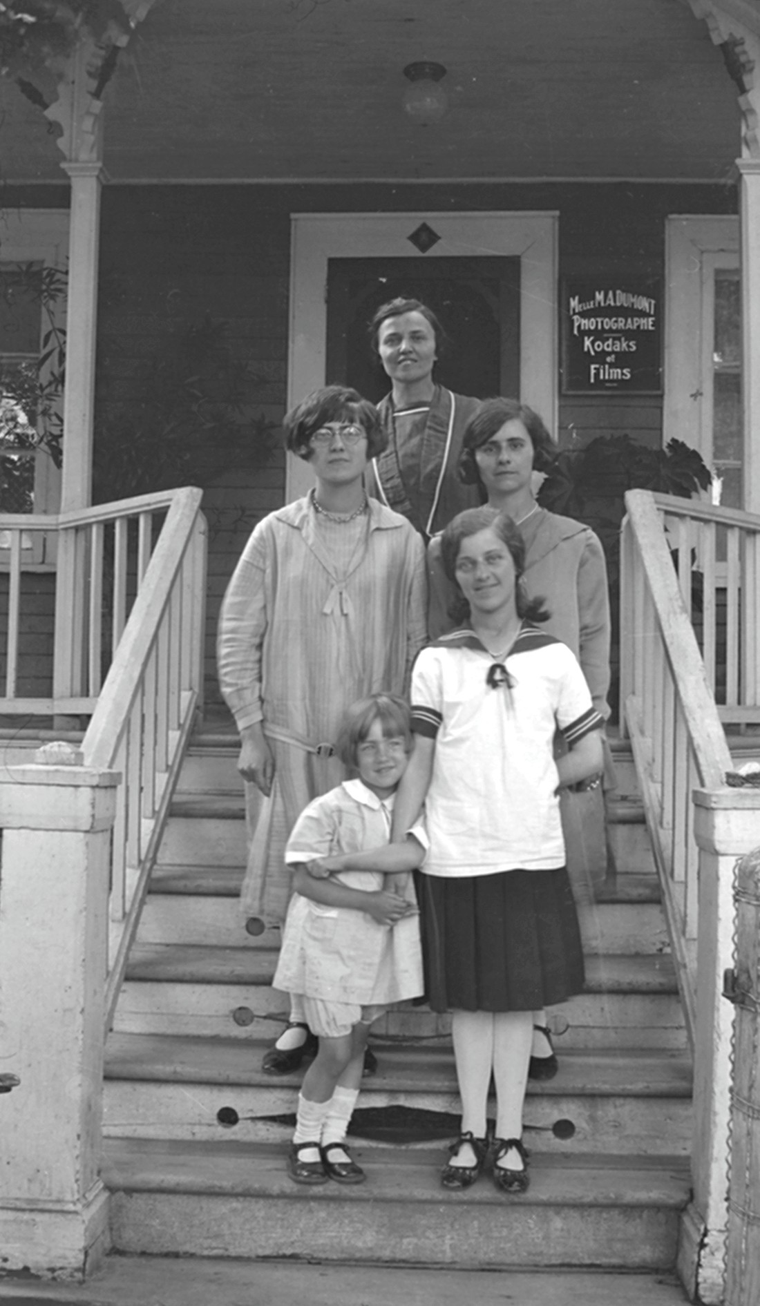 Un groupe de cinq femmes dont une fillette et Marie-Alice Dumont sur le perron du Studio Dumont avec sur la façade de la maison un écriteau sur lequel est inscrit "Melle M.A. Dumont Photographe Kodaks et Films".
