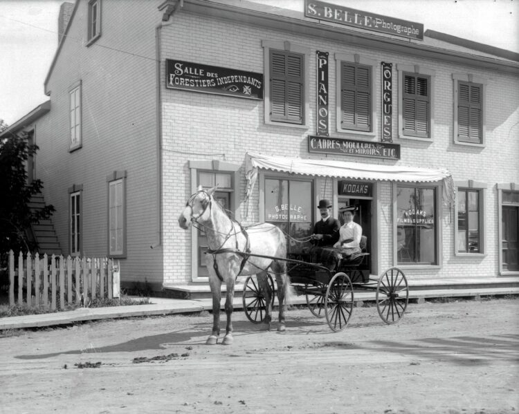 Vue de la façade du studio de Stanislas Belle devant lequel pose un couple assis dans une calèche tirée par un cheval blanc