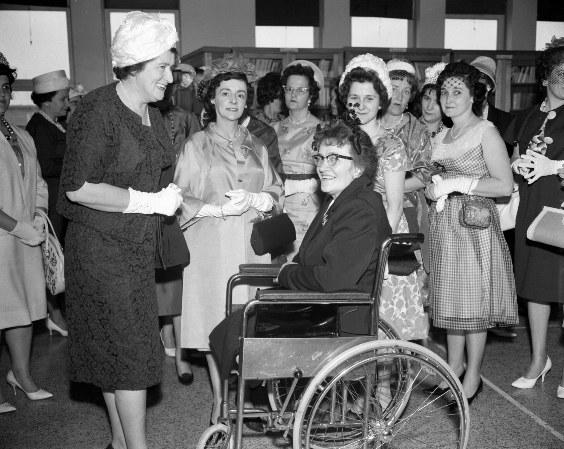 Groupe de femmes élégamment vêtues rassemblées dans une bibliothèque et dont l'une, à l'avant-plan, est en chaise roulante.