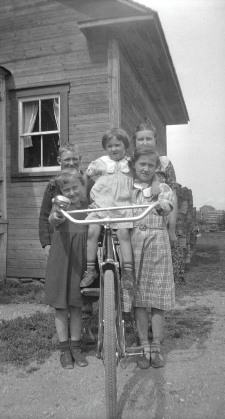 Quatre enfants et une jeune femmes posant avec une bicyclette devant une maison.